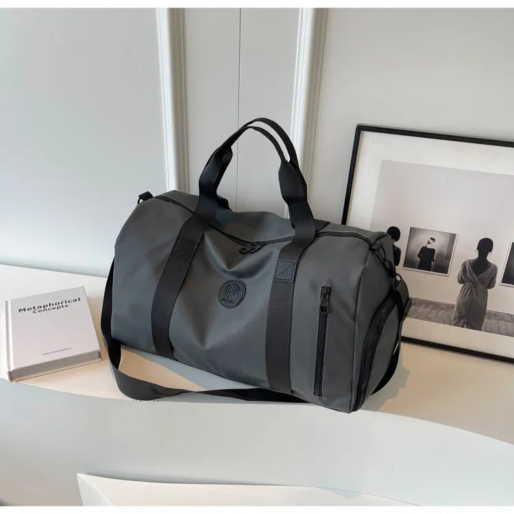 Gray duffel bag with black straps and a circular "BUBM" logo, placed on a white curved surface. Features zippered compartments and padded handles—sleek, minimalist, and ideal for travel or gym use.