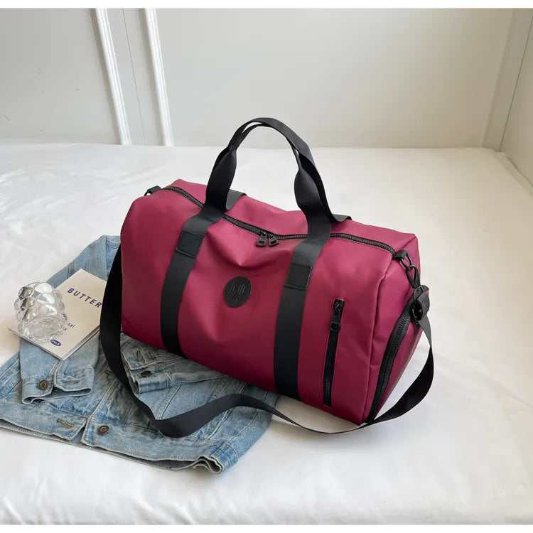 Maroon duffel bag with black straps and a circular logo, placed on a light-colored bed over a blue denim jacket. Nearby, a book titled "BUTTER" and a clear plastic item add a cozy, organized vibe—perfect for travel or casual use.