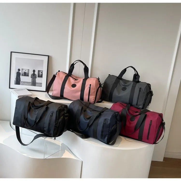 Five cylindrical duffel bags in pink, gray, black, navy blue, and maroon are neatly displayed on a curved white surface in a minimalistic room. Each bag features a circular logo and zippered pocket—stylish and versatile, ideal for travel or gym use.