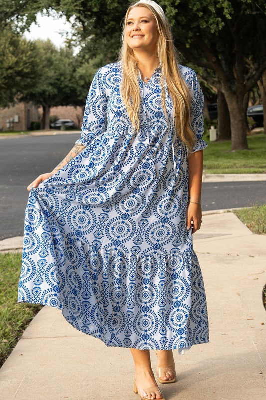 A woman standing on a sidewalk, wearing a blue and white patterned dress with a collar and elbow-length sleeves.