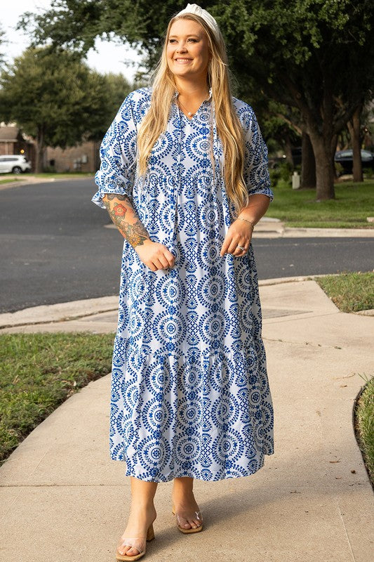 A woman standing on a sidewalk, wearing a blue and white patterned dress with a collar and elbow-length sleeves.
