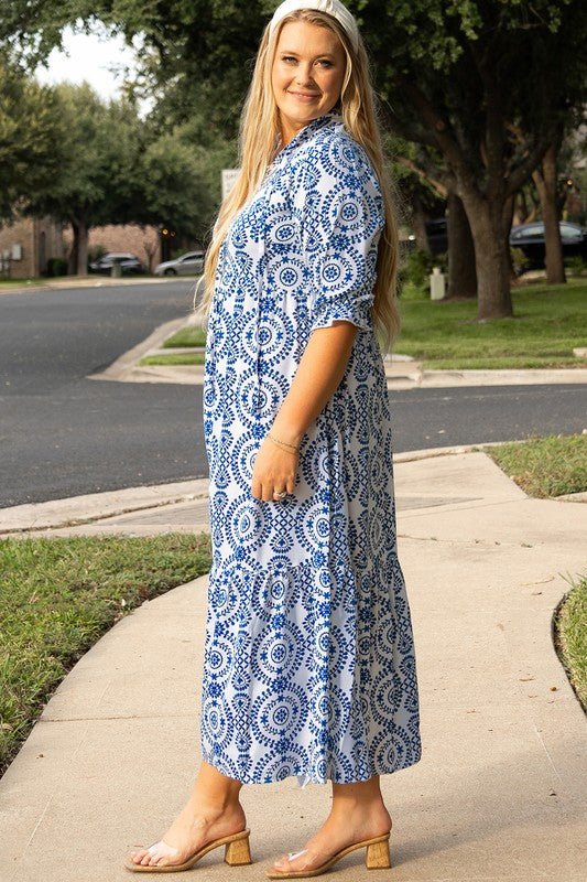 A woman standing on a sidewalk, wearing a blue and white patterned dress with a collar and elbow-length sleeves.