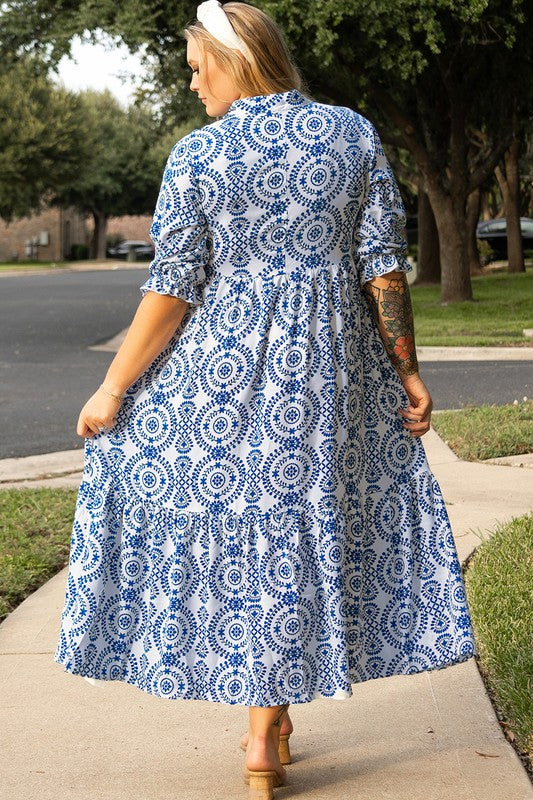 A woman standing on a sidewalk, wearing a blue and white patterned dress with a collar and elbow-length sleeves.