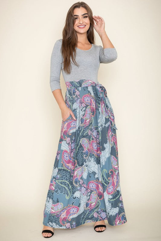 A woman with long brown hair is posing in a stylish outfit. She's wearing a white top with black stripes and a long white skirt with colorful circular patterns. She's got on tan heels and is touching her hair with one hand.