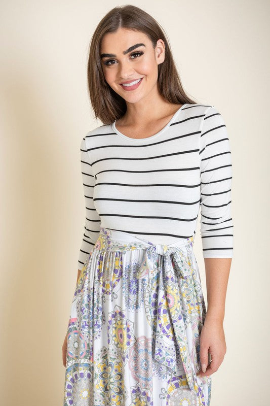 A woman with long brown hair is posing in a stylish outfit. She's wearing a white top with black stripes and a long white skirt with colorful circular patterns. She's got on tan heels and is touching her hair with one hand.
