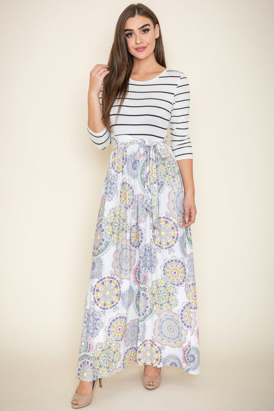 A woman with long brown hair is posing in a stylish outfit. She's wearing a white top with black stripes and a long white skirt with colorful circular patterns. She's got on tan heels and is touching her hair with one hand.