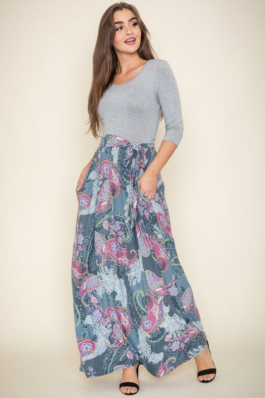 A woman with long brown hair is posing in a stylish outfit. She's wearing a white top with black stripes and a long white skirt with colorful circular patterns. She's got on tan heels and is touching her hair with one hand.