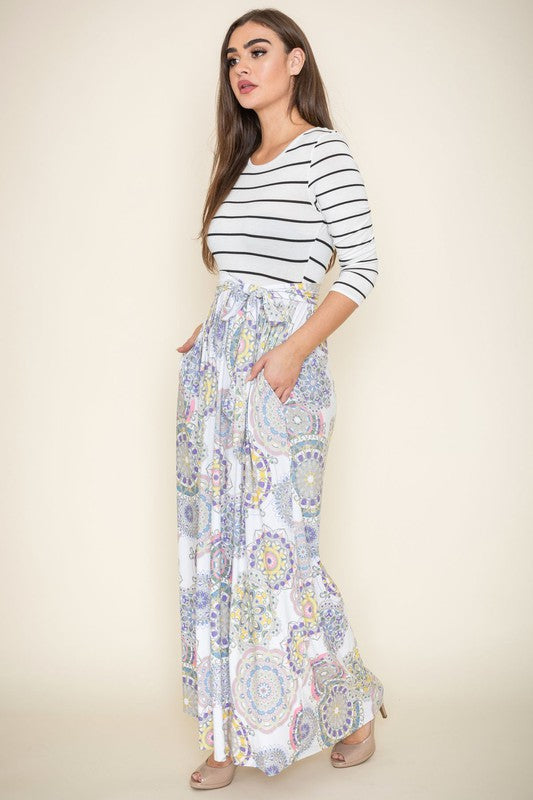 A woman with long brown hair is posing in a stylish outfit. She's wearing a white top with black stripes and a long white skirt with colorful circular patterns. She's got on tan heels and is touching her hair with one hand.
