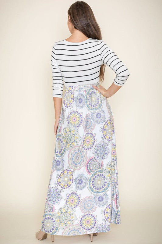 A woman with long brown hair is posing in a stylish outfit. She's wearing a white top with black stripes and a long white skirt with colorful circular patterns. She's got on tan heels and is touching her hair with one hand.