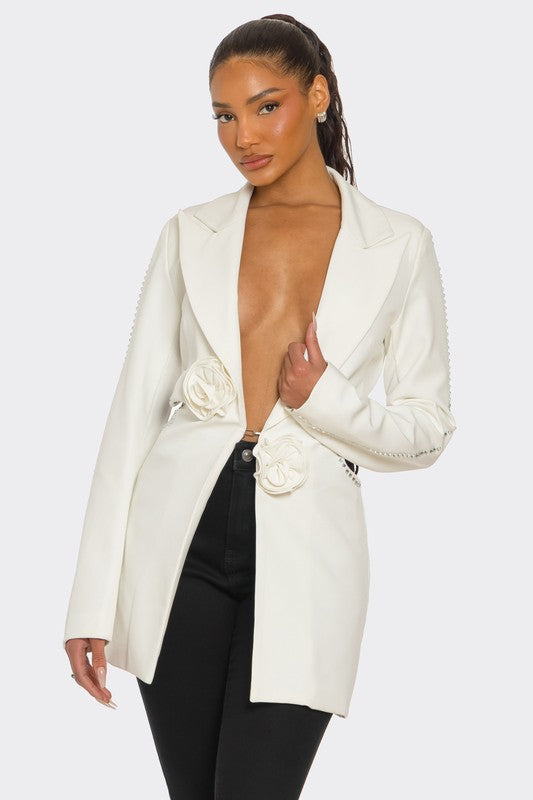 A lady posing in a sleek white blazer with floral details and a daring deep V-neckline, paired with black pants.