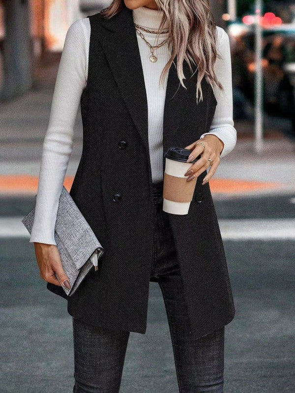 A woman dressed in a stylish outfit, standing on a street. She's wearing a long black vest over a white long-sleeve top and dark jeans. She's holding a coffee cup in one hand and a clutch in the other.