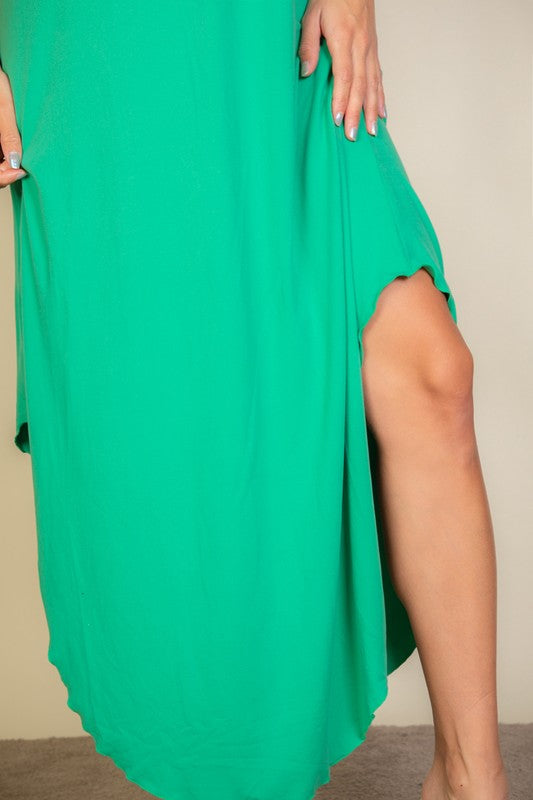 A woman with long brown hair is posing in a green, sleeveless midi dress with a V-neck and a slit on one side. She's wearing tan heels and standing on a beige carpet against a beige wall.