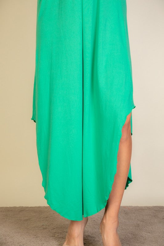 A woman with long brown hair is posing in a green, sleeveless midi dress with a V-neck and a slit on one side. She's wearing tan heels and standing on a beige carpet against a beige wall.