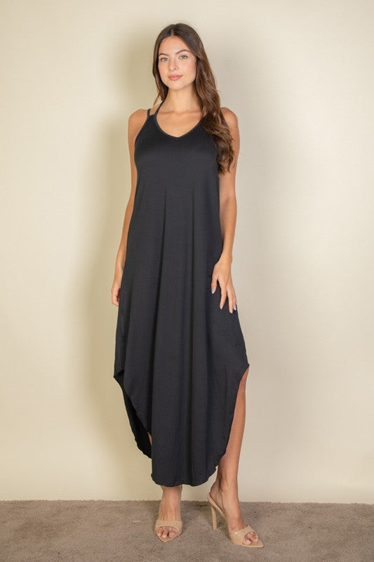 A woman with long brown hair is posing in a black, sleeveless midi dress with a V-neck and a slit on one side. She's wearing tan heels and standing on a beige carpet against a beige wall.