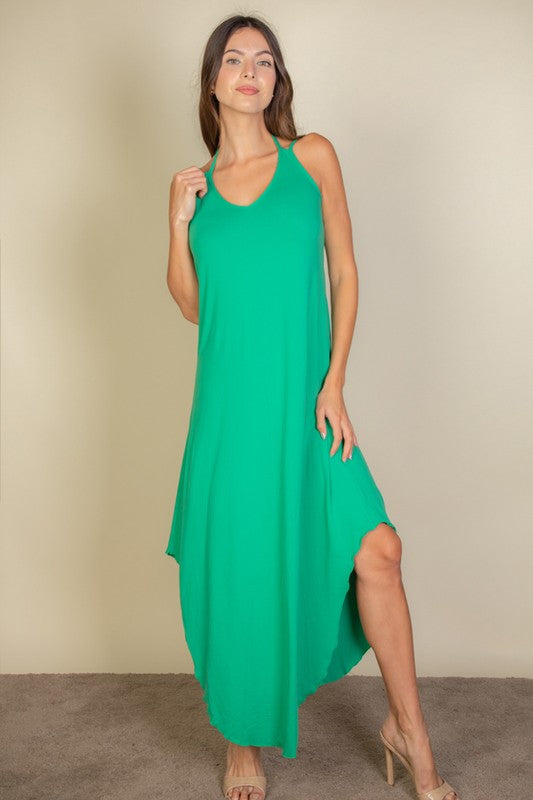 A woman with long brown hair is posing in a green, sleeveless midi dress with a V-neck and a slit on one side. She's wearing tan heels and standing on a beige carpet against a beige wall