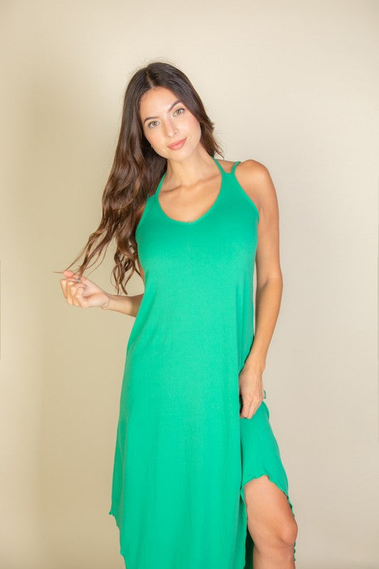 A woman with long brown hair is posing in a green, sleeveless midi dress with a V-neck and a slit on one side. She's wearing tan heels and standing on a beige carpet against a beige wall.