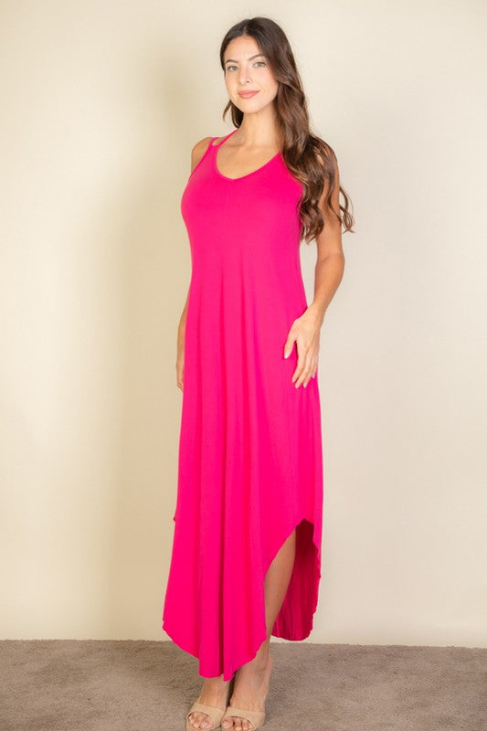 A woman with long brown hair is posing in a pink, sleeveless midi dress with a V-neck and a slit on one side. She's wearing tan heels and standing on a beige carpet against a beige wall.