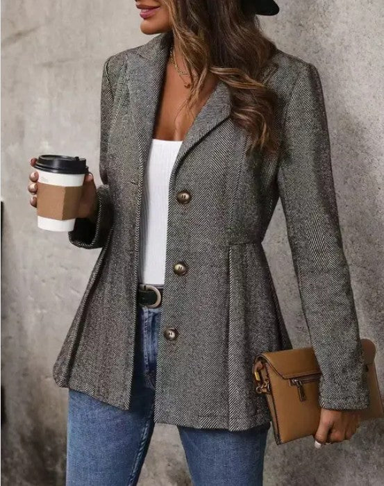 A woman wearing a gray tweed blazer with brass buttons, a white top, blue jeans, and a black hat. She's holding a coffee cup in one hand and a brown purse in the other, posing against a gray wall.
