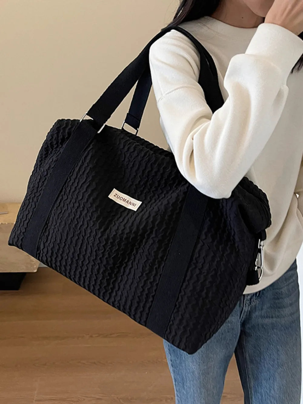 A woman in a white long-sleeve top and blue jeans carries a large black quilted duffel bag over her shoulder. She stands indoors on a wooden floor near a wooden bench—casual and stylish look.