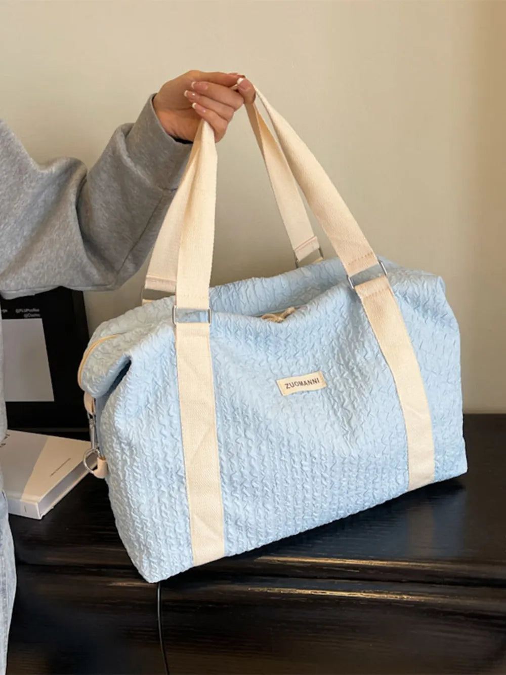Light blue quilted duffel bag with beige straps, held by a person in a grey long-sleeve top. The bag features a front label and rests on a dark wooden surface—soft, stylish, and practical.