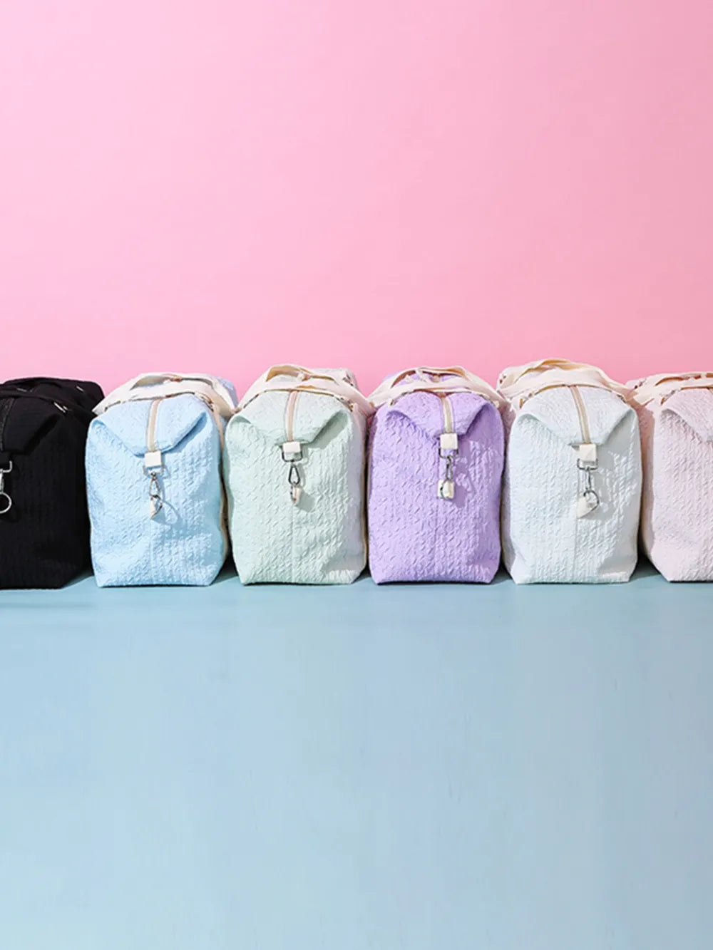 A row of seven quilted backpacks in various colors—black, light blue, mint green, lavender, white, and light pink—neatly arranged against a pastel pink and blue background. Uniform design with flap closures and top handles—stylish product lineup.