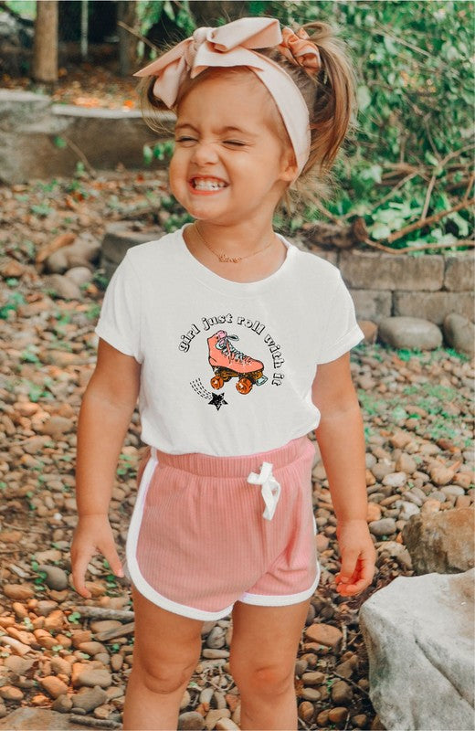 Child wearing a white t-shirt with a graphic and pink shorts outdoors on a rocky ground.