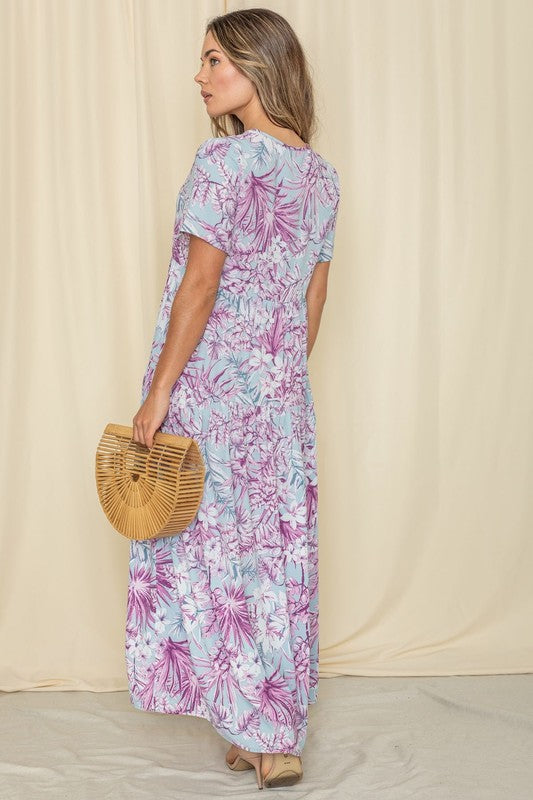 A woman in a floral maxi dress with short sleeves, standing against a cream curtain backdrop.
