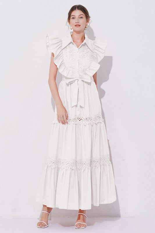Woman wearing a sleeveless embroidered maxi dress with dramatic ruffle sleeves, tiered lace skirt, collared neckline, and waist tie, styled with heels.