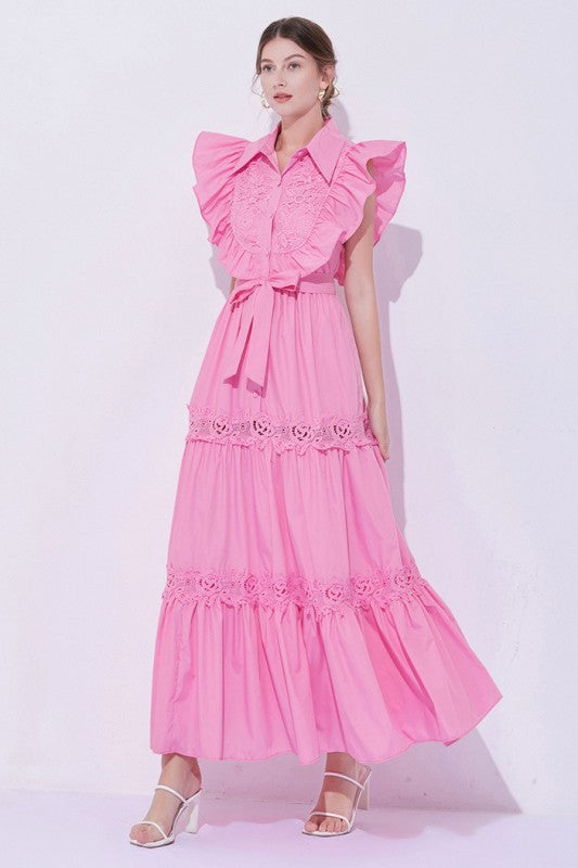 Woman wearing a sleeveless embroidered maxi dress with dramatic ruffle sleeves, tiered lace skirt, collared neckline, and waist tie, styled with heels.