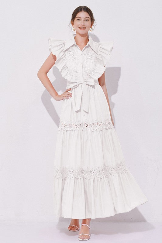 Woman wearing a sleeveless embroidered maxi dress with dramatic ruffle sleeves, tiered lace skirt, collared neckline, and waist tie, styled with heels.