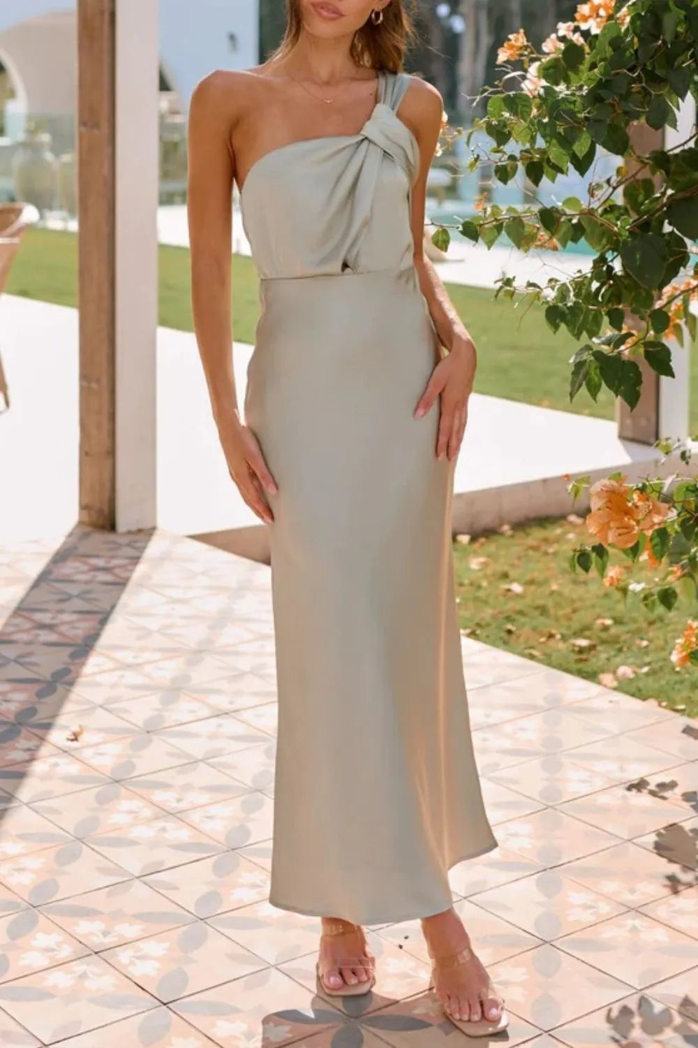 A light taupe, satin-like, sleeveless maxi dress with a draped, twisted front detail and a sleek, form-fitting silhouette.