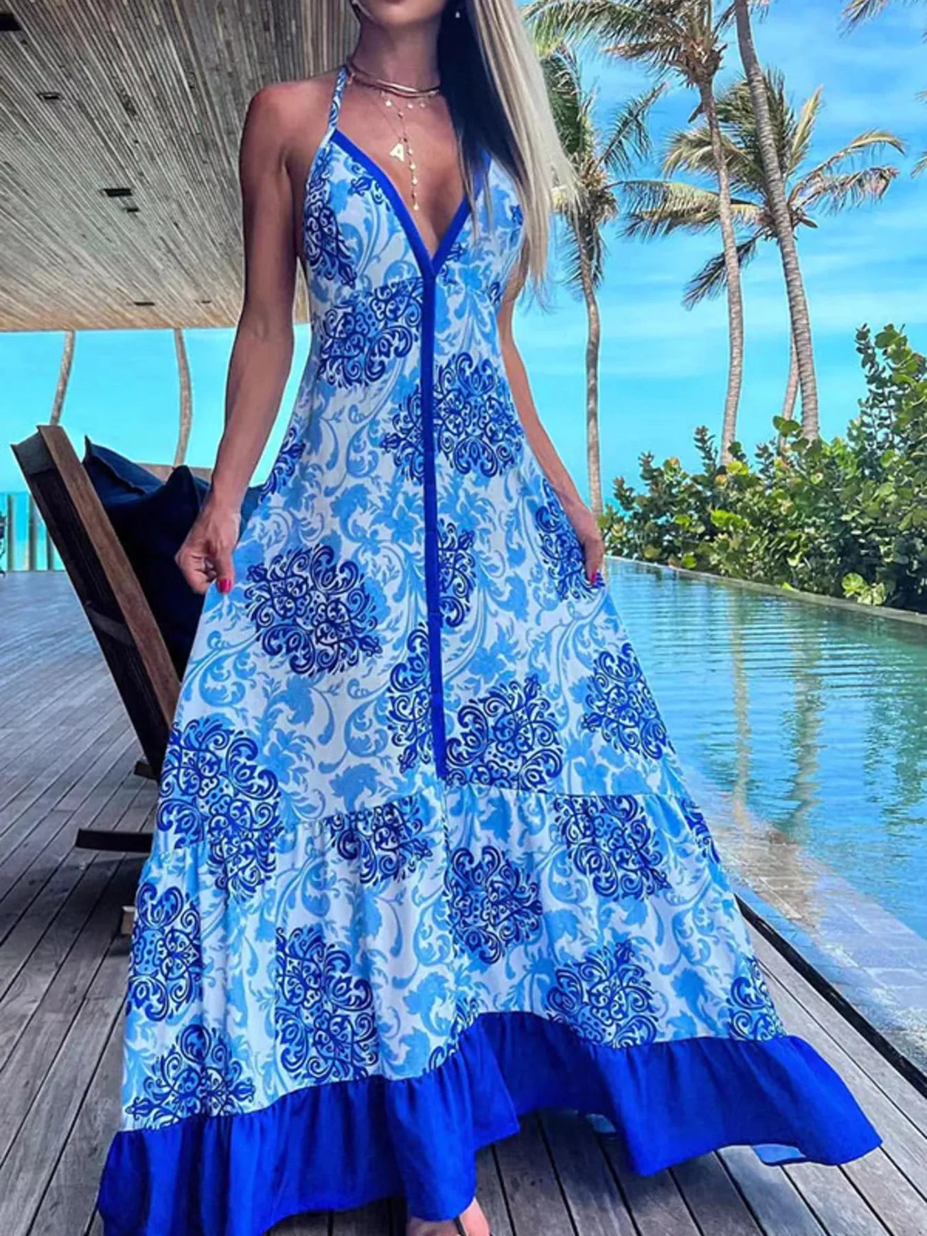 Woman wearing a blue and white floral halter-neck maxi dress with deep V-neckline and solid trim, standing by a tropical infinity pool