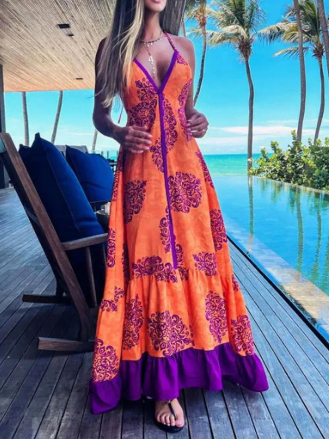 Woman wearing a floral halter-neck maxi dress with deep V-neckline and solid trim, standing by a tropical infinity pool