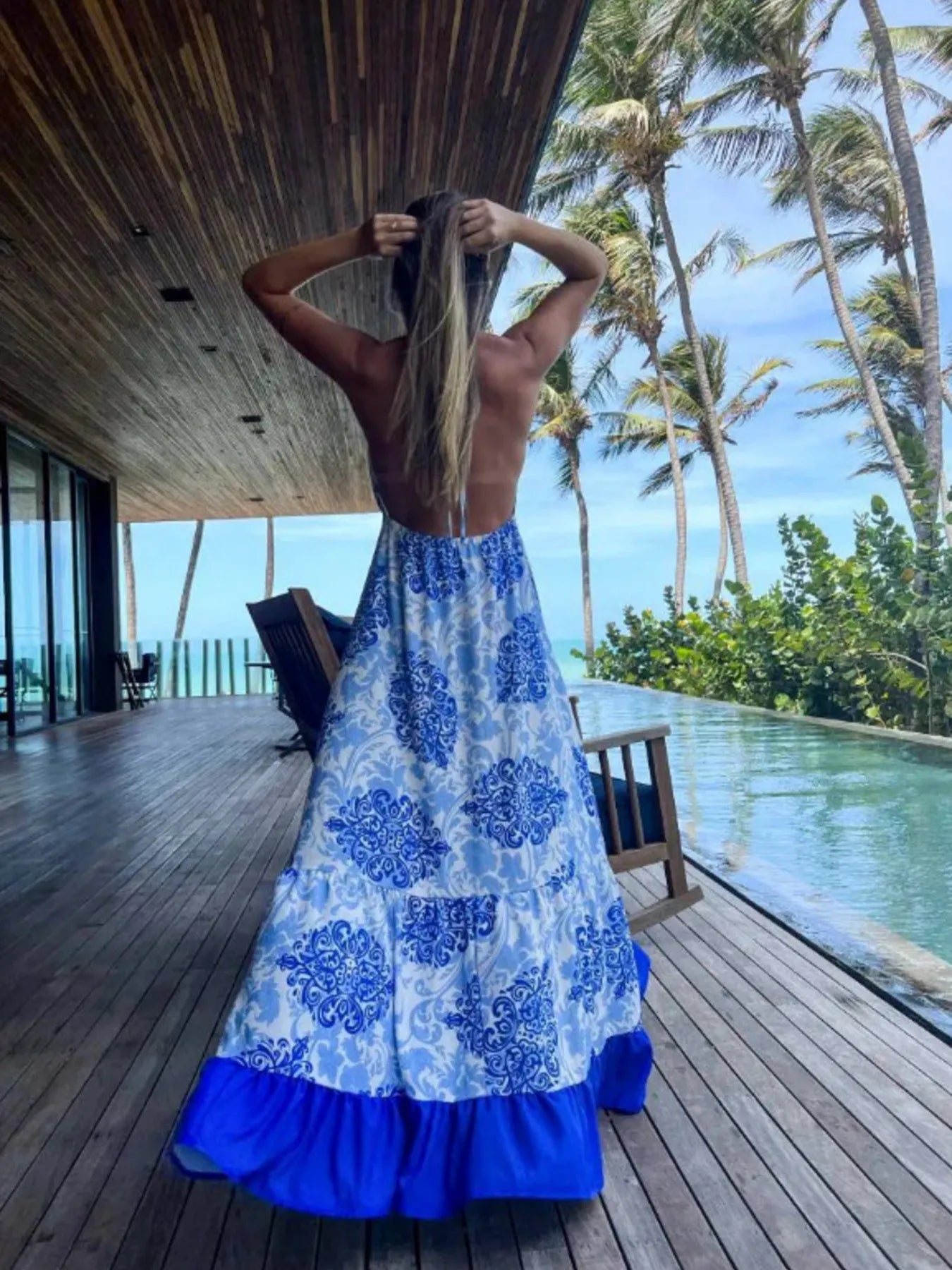 Woman wearing a blue and white floral halter-neck maxi dress with deep V-neckline and solid trim, standing by a tropical infinity pool