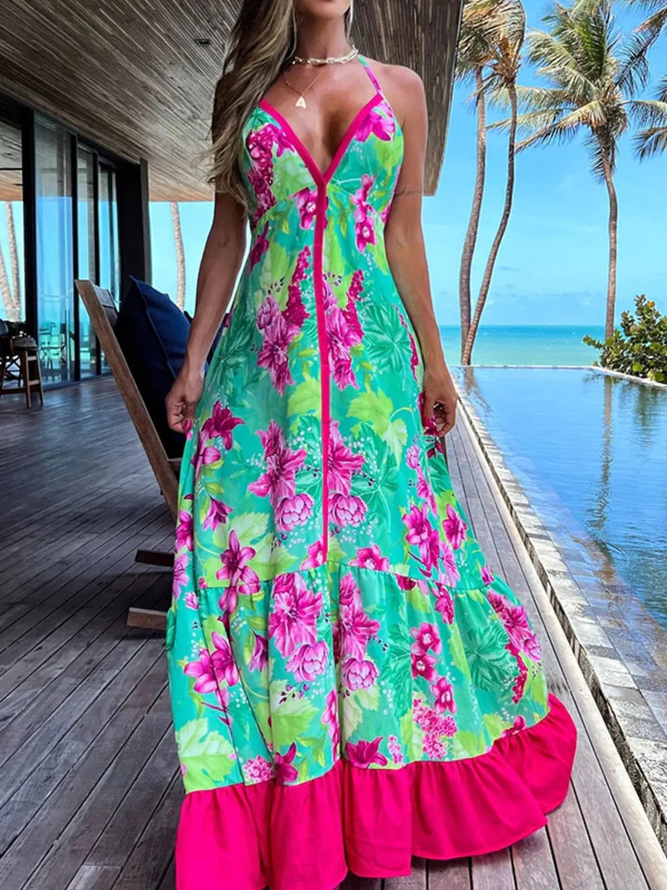 Woman wearing a floral halter-neck maxi dress with deep V-neckline and solid trim, standing by a tropical infinity pool