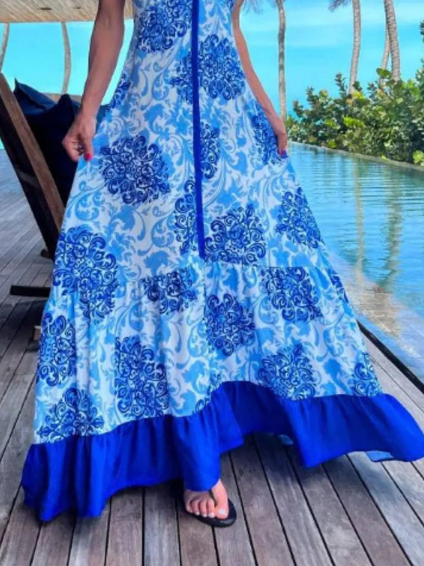 Woman wearing a blue and white floral halter-neck maxi dress with deep V-neckline and solid trim, standing by a tropical infinity pool