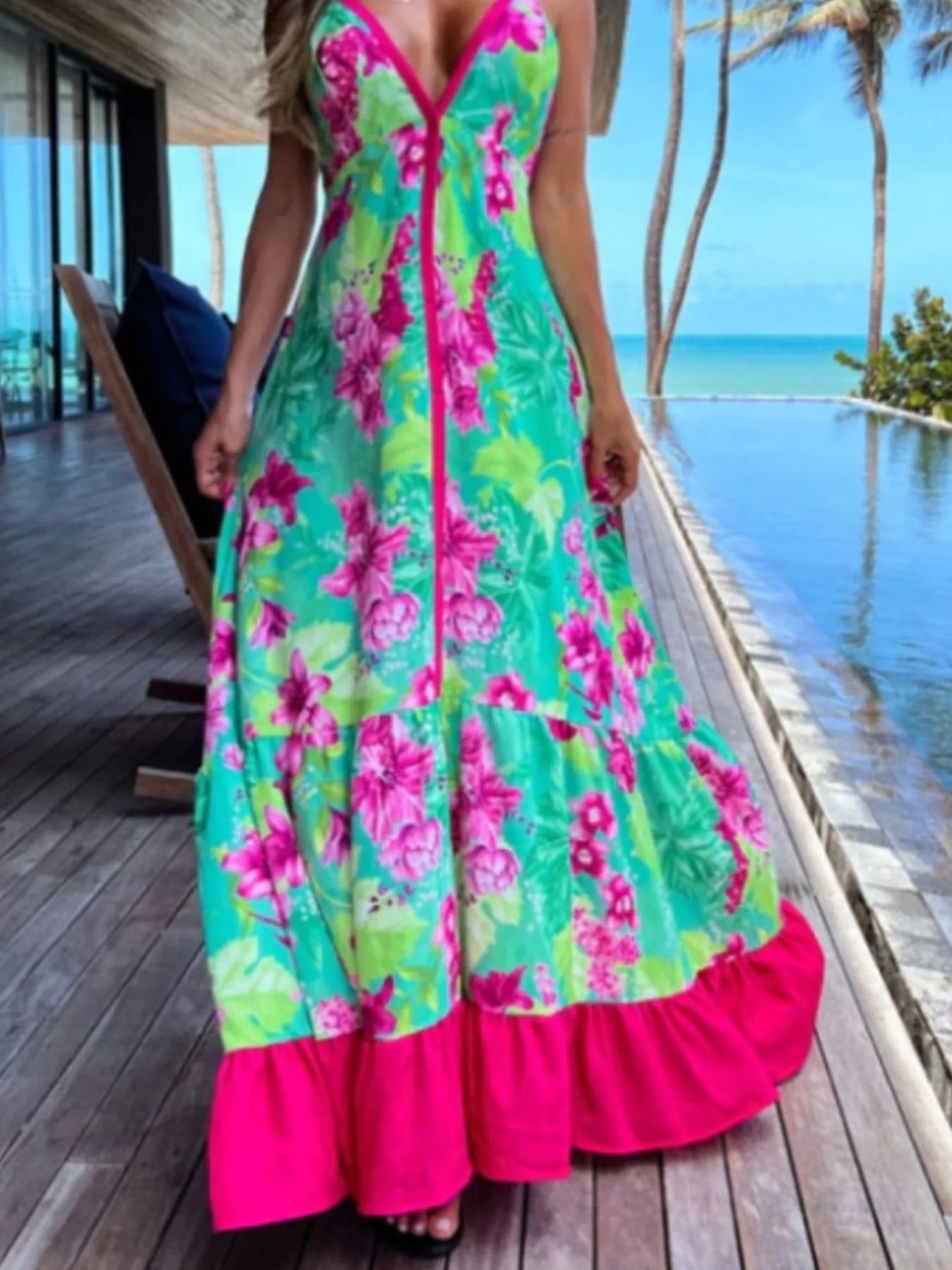 Woman wearing a floral halter-neck maxi dress with deep V-neckline and solid trim, standing by a tropical infinity pool
