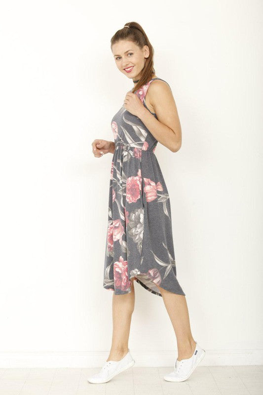 Woman poses in a stylish sleeveless floral midi dress with pink flower print, featuring a flattering gathered waist and practical pockets, full view.