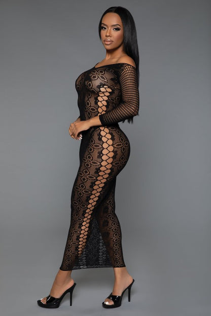 The dress is a form-fitting, full-length black lace bodysuit with long sleeves and a sheer, intricate pattern that reveals the silhouette underneath, paired with open-toe heels for a sultry, elegant look.