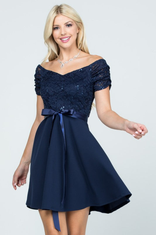 The dress is a knee-length, navy blue frock with a fitted, off-the-shoulder bodice adorned with delicate lace or textured fabric and a full, flowing skirt tied at the waist with a matching sash.