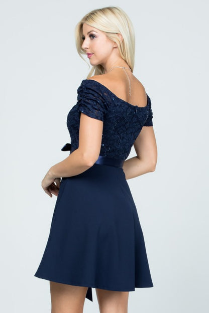 The dress is a knee-length, navy blue frock with a fitted, off-the-shoulder bodice adorned with delicate lace or textured fabric and a full, flowing skirt tied at the waist with a matching sash.