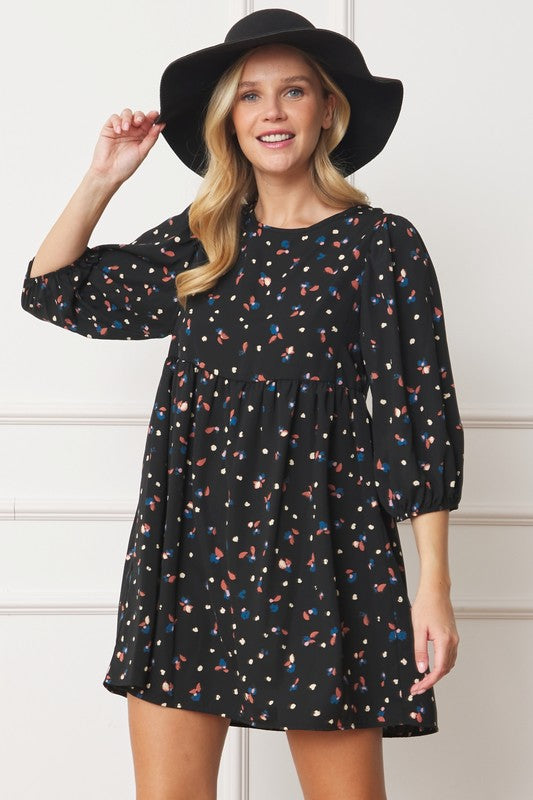 The dress is a short, loose-fitting black shift adorned with a delicate, colorful floral print and three-quarter sleeves