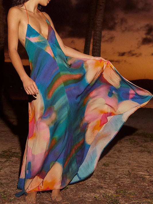 Woman wearing a flowing abstract print halter-neck maxi cami dress with deep neckline, standing outdoors at sunset with palm trees in the background.
