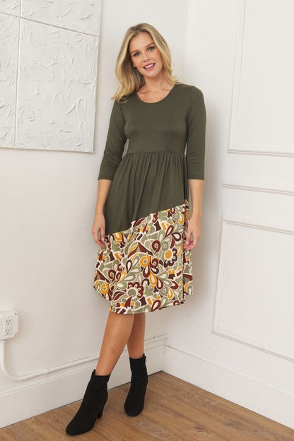 Woman wearing an olive green midi dress with a colorful, abstract floral or paisley pattern on the bottom half, full shot view.