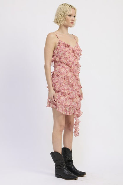 The dress is a pink, asymmetrical, ruffled mini dress with delicate spaghetti straps, featuring a loose, draped silhouette and a slightly sheer, layered texture that gives it a playful, feminine look.
