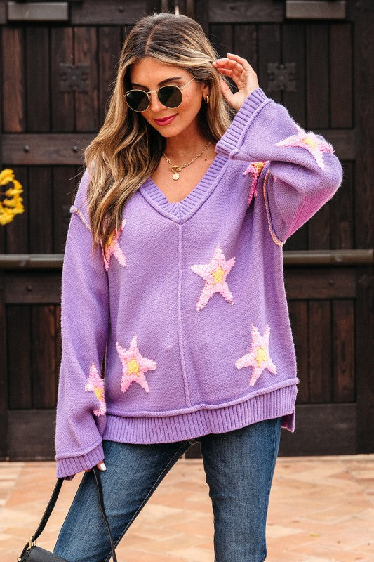 Woman wearing a purple oversized V-neck sweater with pink and yellow star patches, with fingers running through hair.