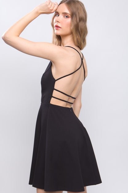 the dress is a light beige, knee-length A-line with a delicate back design featuring thin crisscross straps that wrap around the back, giving a soft, feminine silhouette.