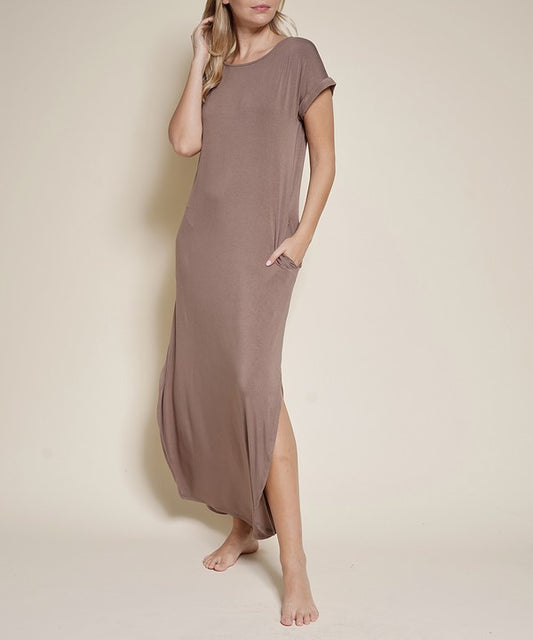 The image shows a woman wearing a comfy-looking brown maxi dress with short sleeves and a pocket on the side. She's barefoot, posing casually with one hand in her pocket and the other touching her hair against a plain beige background. Slaying the effortless vibe.