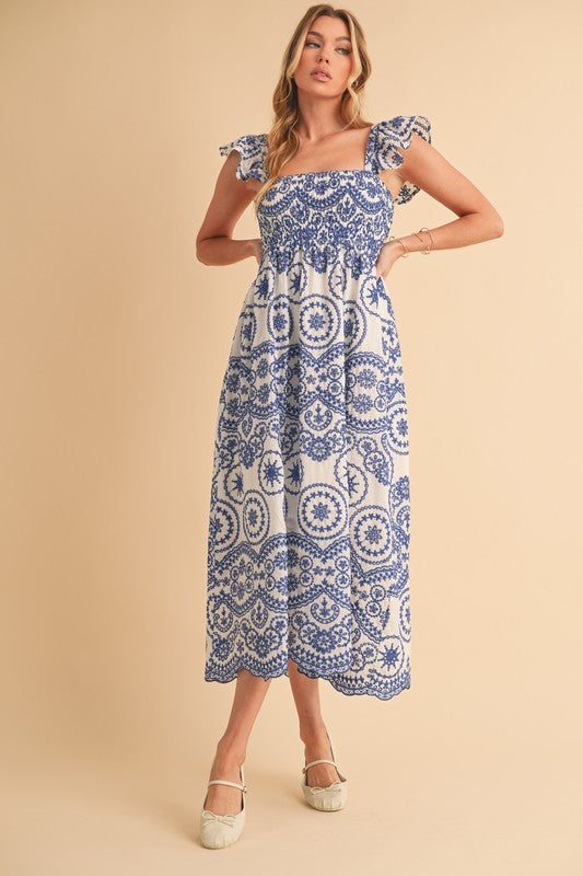 The dress is a long, airy maxi with a blue and white geometric/medallion print, cap sleeves, and a fitted bodice that flows into a relaxed, ankle-length skirt.