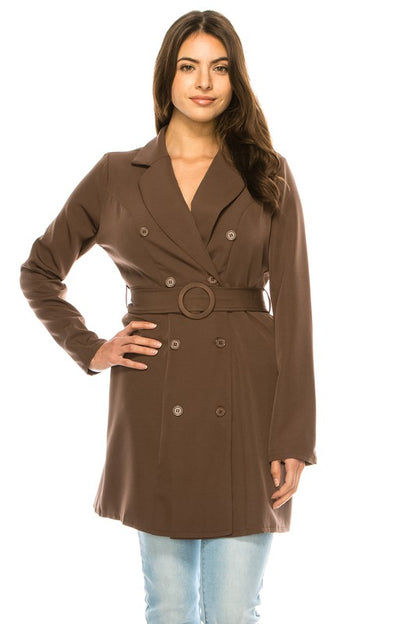 A woman wearing a brown double-breasted coat with a belt and light blue jeans. She's standing against a white background.